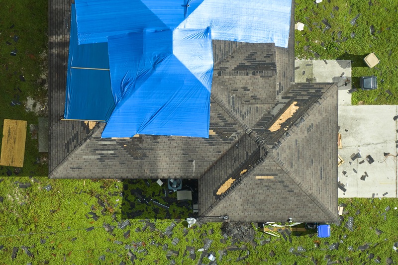Top view of a house with blue tarps on the roof. Shingles and debris are scattered all around, indicating damage after a storm or high winds.